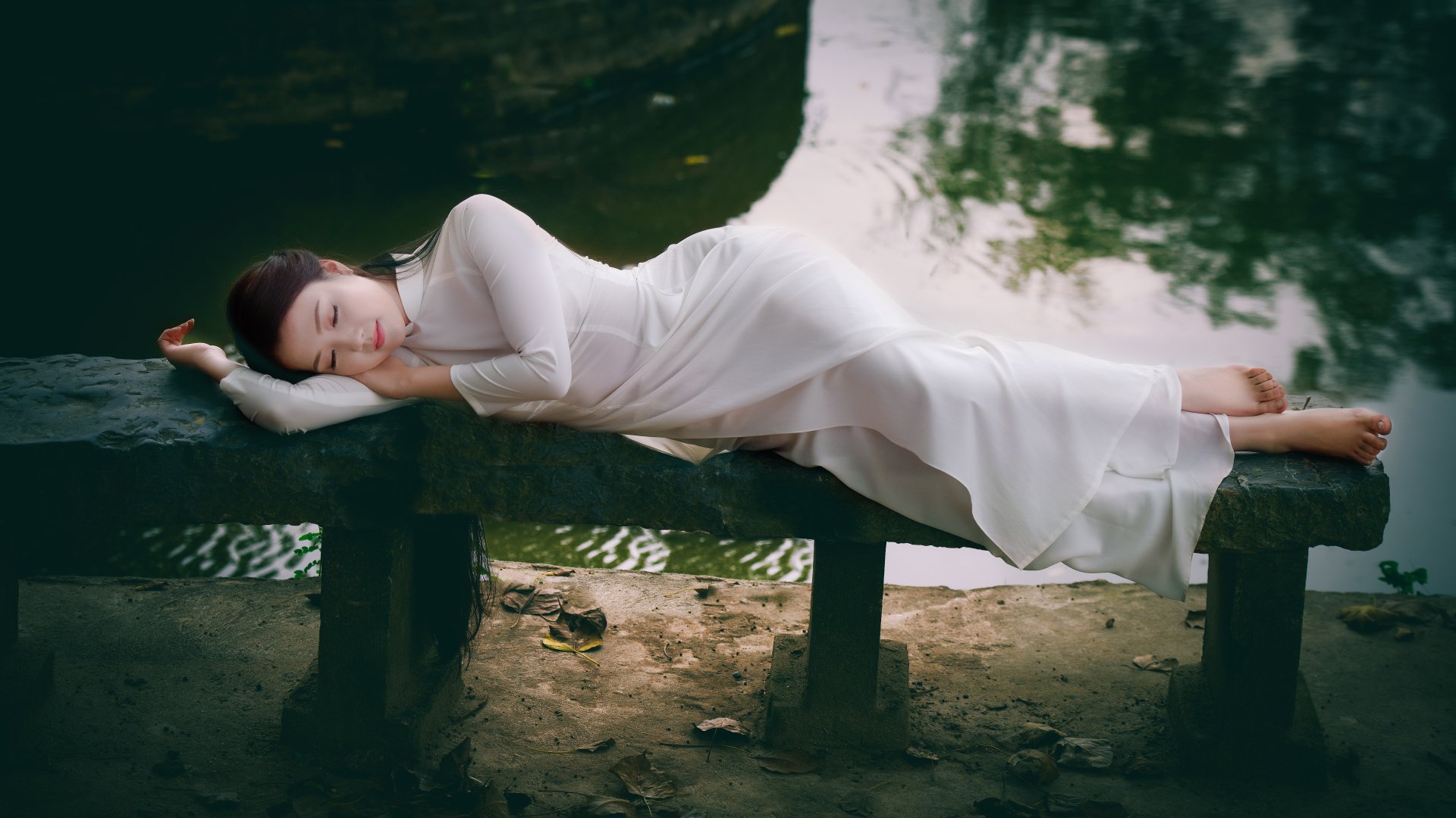 A Vietnamese woman in an Ao Dai lies peacefully on a bench by a serene pond, creating a tranquil and picturesque HD desktop wallpaper and background.