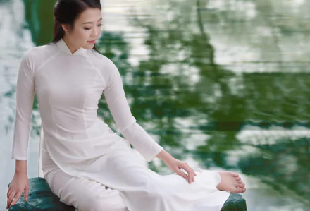 A Vietnamese woman in a traditional white Ao Dai sits gracefully by calm water, captured in a serene 4K Ultra HD desktop wallpaper.