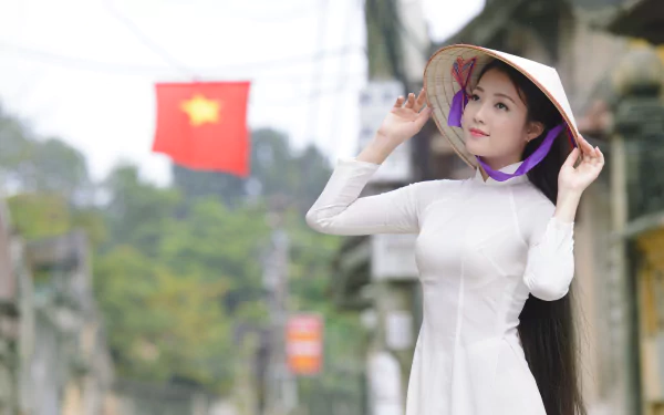 Vietnamese woman wearing a traditional Ao Dai and Asian conical hat, standing outdoors with a blurred Vietnamese flag in the background, captured in 4K Ultra HD.