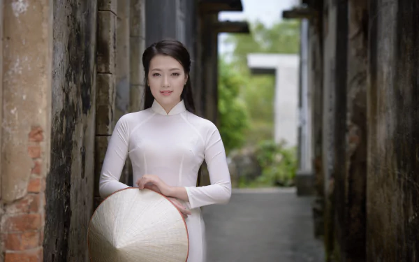 Vietnamese woman in a traditional white Ao Dai holding an Asian conical hat, standing between weathered pillars in a serene outdoor setting, captured in 4K Ultra HD.