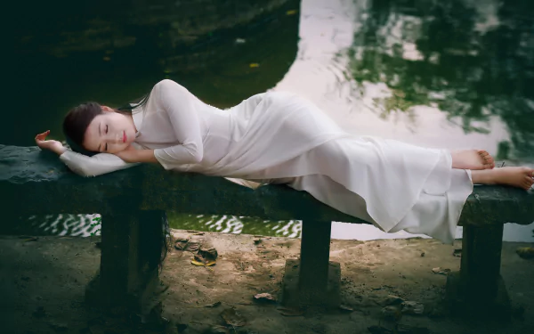A Vietnamese woman in an Ao Dai lies peacefully on a bench by a serene pond, creating a tranquil and picturesque HD desktop wallpaper and background.