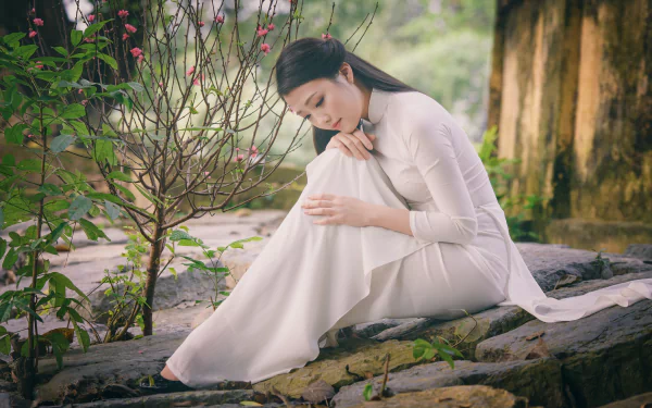 A Vietnamese woman in a white Ao Dai sits gracefully on a rock beside blooming spring flowers. The serene setting and high-definition quality make this a beautiful HD desktop wallpaper and background.