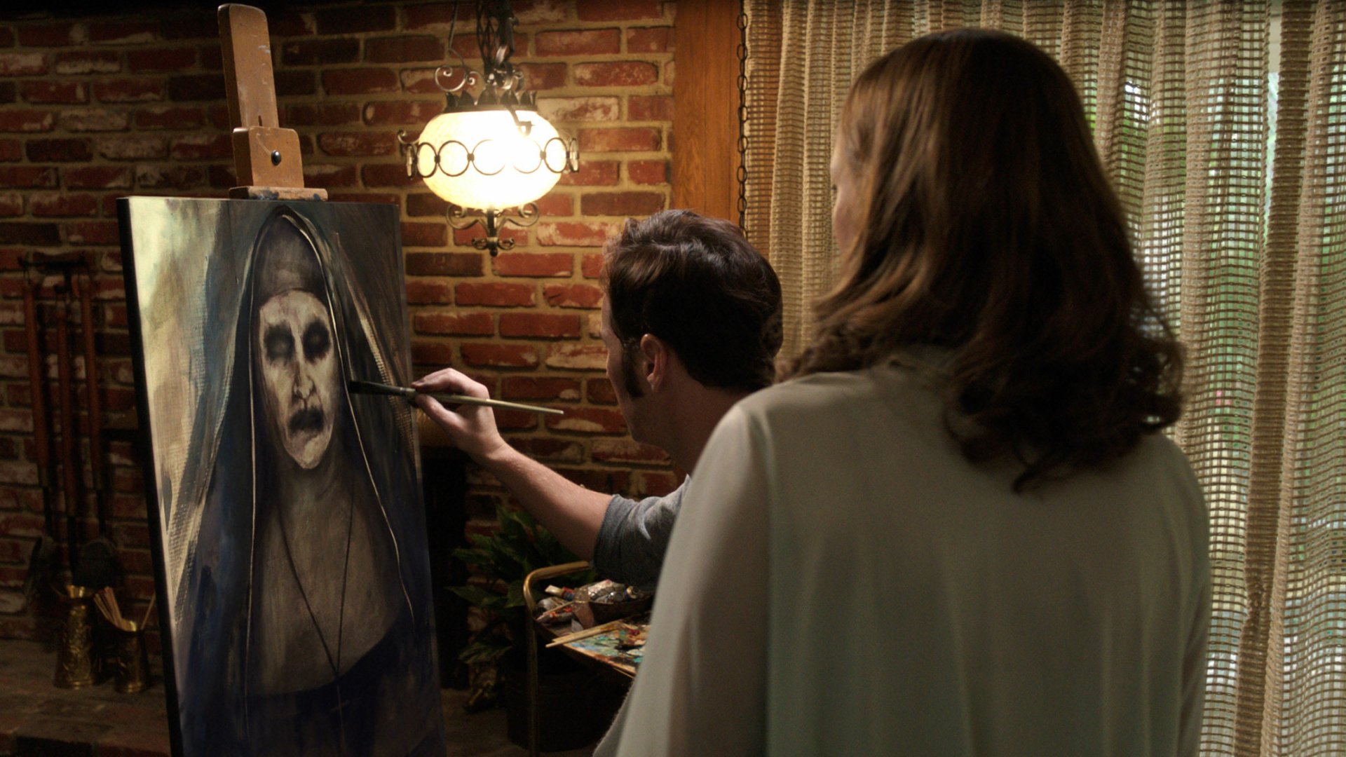 A scene from *The Conjuring 2* shows a man painting a haunting portrait of a nun, while a woman observes. The rustic setting enhances the eerie atmosphere of the film.