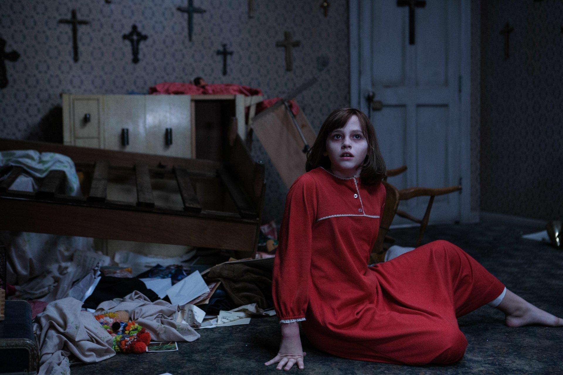 A tense scene from *The Conjuring 2*, featuring Sterling Jerins in a red nightgown, sitting on a messy floor surrounded by debris and crosses, evoking a haunting atmosphere.