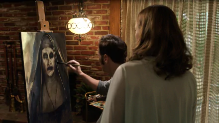 A scene from *The Conjuring 2* shows a man painting a haunting portrait of a nun, while a woman observes. The rustic setting enhances the eerie atmosphere of the film.