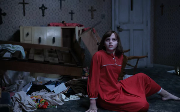 A tense scene from *The Conjuring 2*, featuring Sterling Jerins in a red nightgown, sitting on a messy floor surrounded by debris and crosses, evoking a haunting atmosphere.