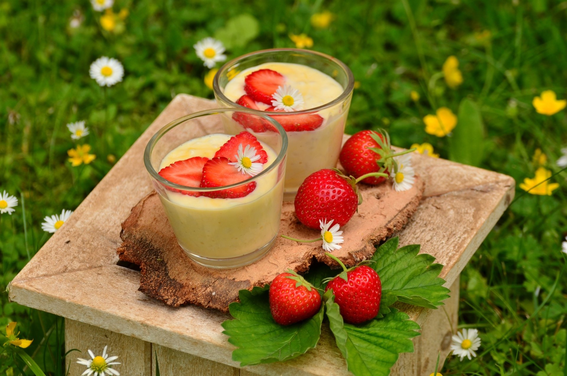 4K Ultra HD wallpaper featuring two glass cups of creamy dessert topped with sliced strawberries and small white flowers, placed on a wooden block amidst green leaves and yellow flowers.