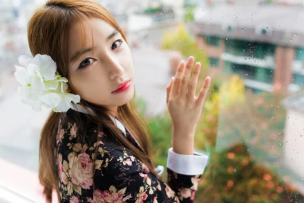 HD wallpaper of an Asian brunette model with brown eyes and lipstick, wearing a floral outfit, posing by a window with a white flower in her hair.