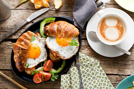 A 4K Ultra HD desktop wallpaper showing a breakfast spread with croissant sandwiches topped with sunny-side-up eggs, fresh vegetables, and a cup of coffee on a rustic wooden table.