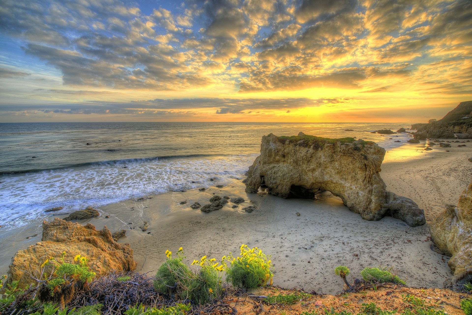 HD desktop wallpaper of a beach at sunset, showcasing the ocean horizon, rocky formations, and vibrant natural colors.