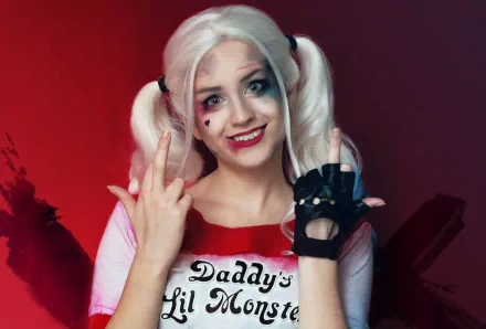 HD PC desktop wallpaper featuring a woman in Harley Quinn Suicide Squad cosplay with blonde pigtails, colorful makeup, and a Daddy's Lil Monster shirt against a red background.