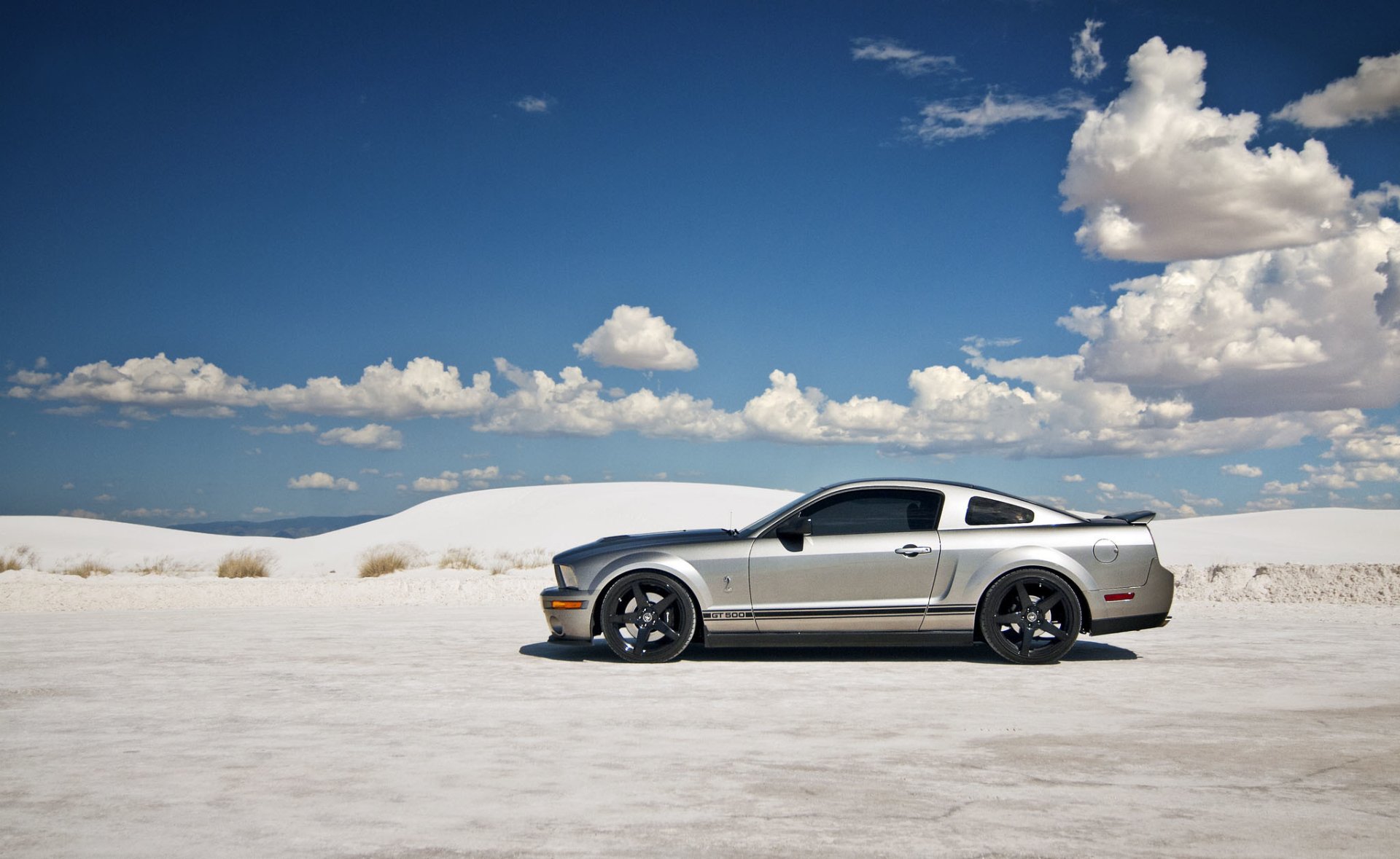 Silver Ford Mustang Shelby GT500 muscle car on white desert salt flats beneath a vivid blue sky with clouds — HD PC desktop wallpaper