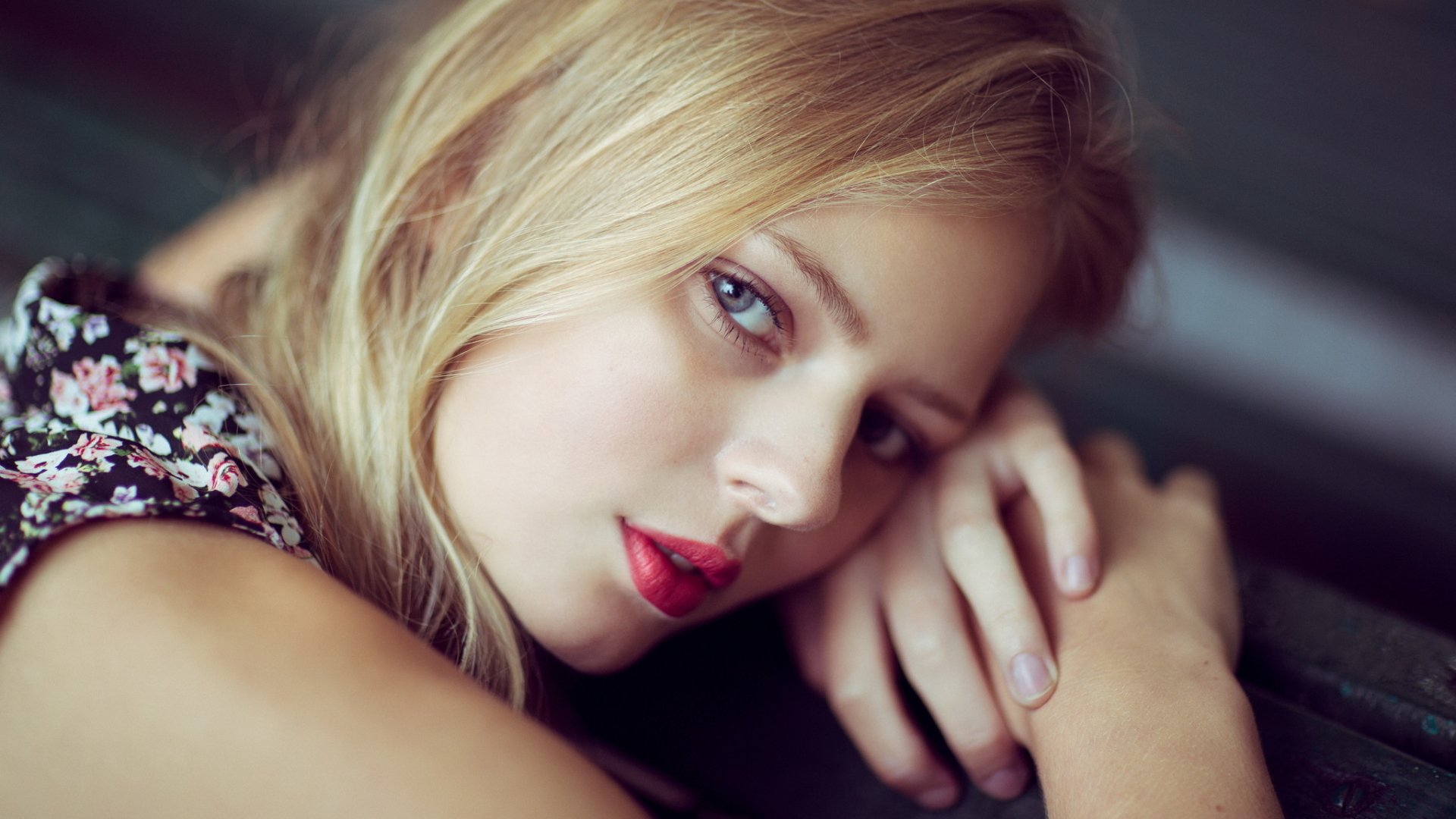 HD wallpaper featuring the portrait of a blonde woman with a serene expression, showcasing her face. The model's hands rest gently on a surface, highlighting her natural beauty.