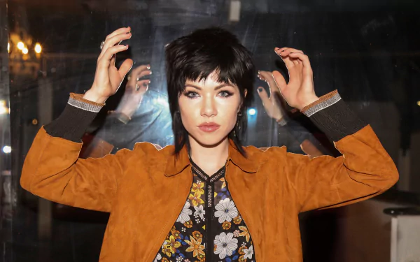 HD desktop wallpaper of a Canadian brunette singer with blue eyes, floral top and brown jacket, hands raised against a reflective studio backdrop — a music-themed portrait.