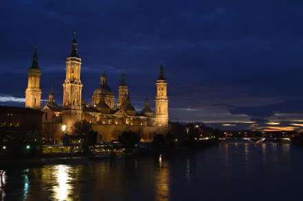 A serene night view of the Basilica of Our Lady of the Pillar in Spain, illuminated alongside a river, showcasing its architectural beauty against the darkening sky.