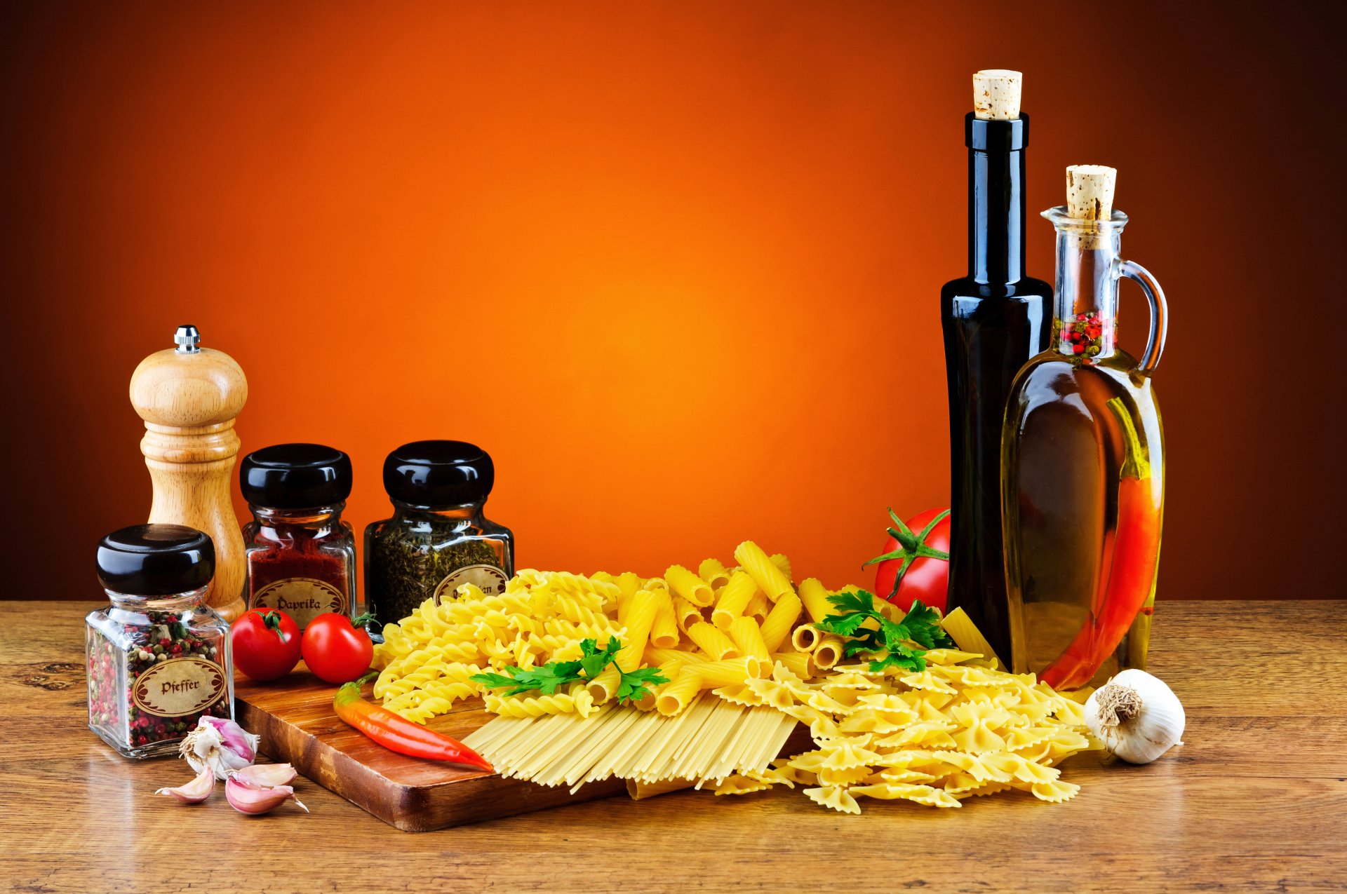 4K Ultra HD still life of various pasta types, olive oil bottles, spices, garlic, and cherry tomatoes arranged on a wooden surface against an orange background.