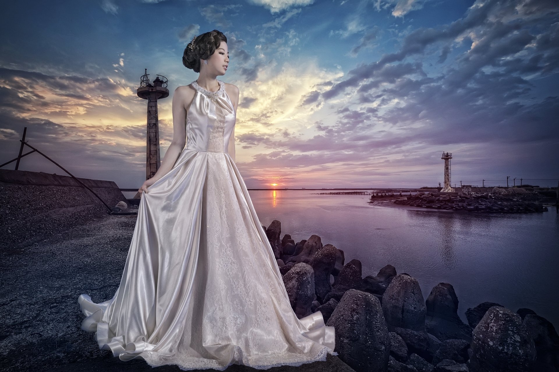 A Chinese woman in a flowing dress stands by the water at sunset, with a dramatic sky and rocky shoreline enhancing the serene atmosphere of the scene.