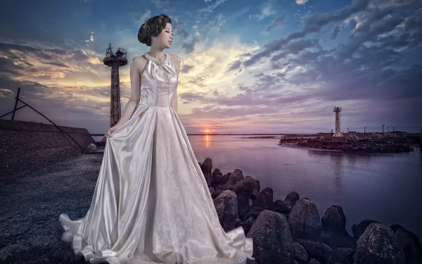 A Chinese woman in a flowing dress stands by the water at sunset, with a dramatic sky and rocky shoreline enhancing the serene atmosphere of the scene.