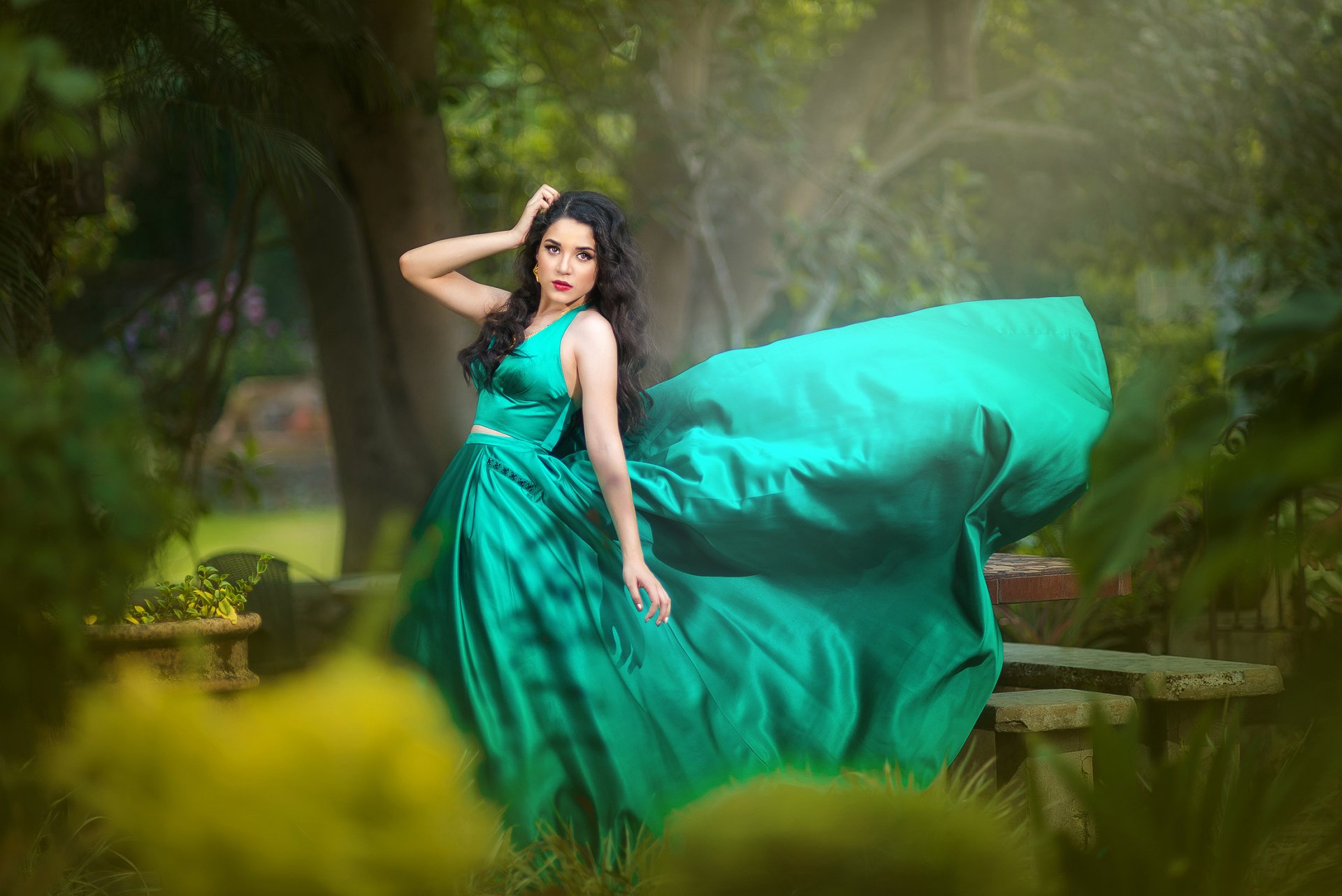A brunette model with brown eyes wearing a flowing green dress and red lipstick poses gracefully outdoors in a lush, green setting.