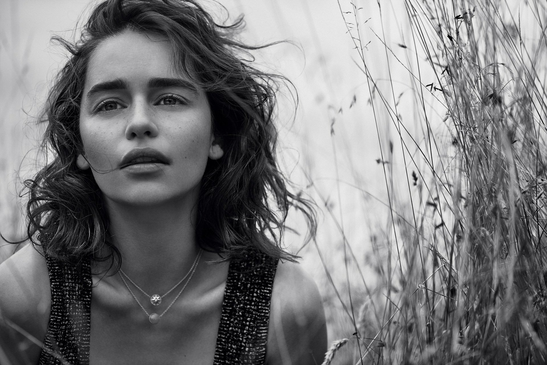 Black and white HD desktop wallpaper featuring celebrity Emilia Clarke with tousled hair, wearing layered necklaces, standing amidst tall grasses.