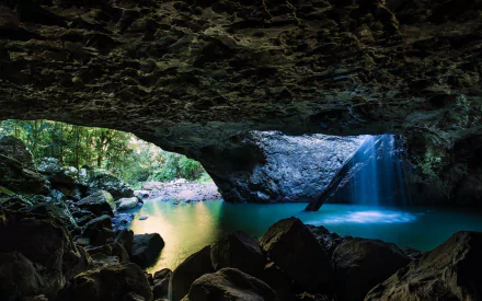 2K Quad HD PC desktop wallpaper and background showing a nature cave—sunlit opening to a turquoise pool with a waterfall and lush greenery beyond.