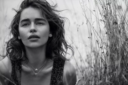 Black and white HD desktop wallpaper featuring celebrity Emilia Clarke with tousled hair, wearing layered necklaces, standing amidst tall grasses.
