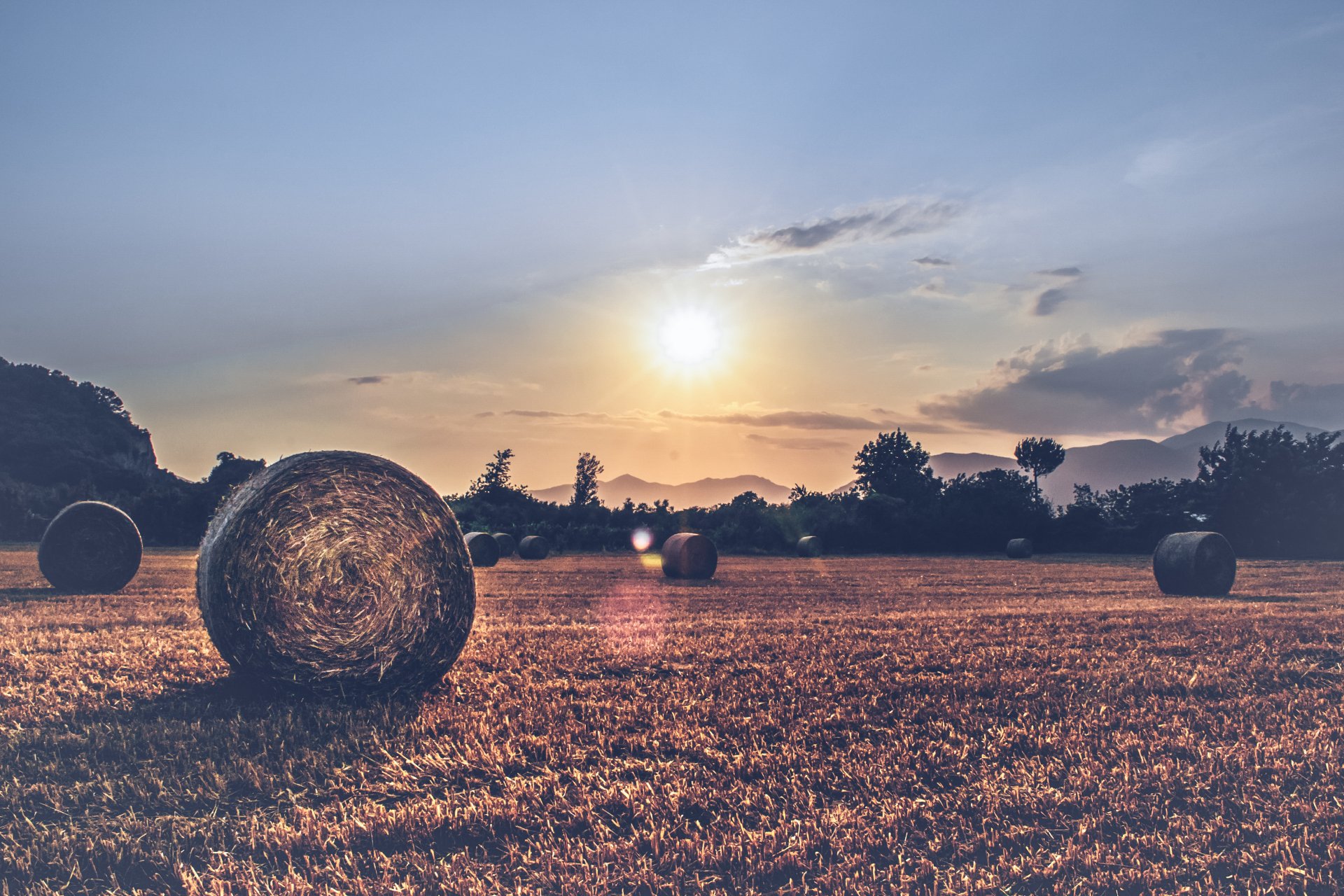 A 4K Ultra HD desktop wallpaper of a sunlit field with haystacks scattered across the landscape under a partly cloudy sky at sunset.