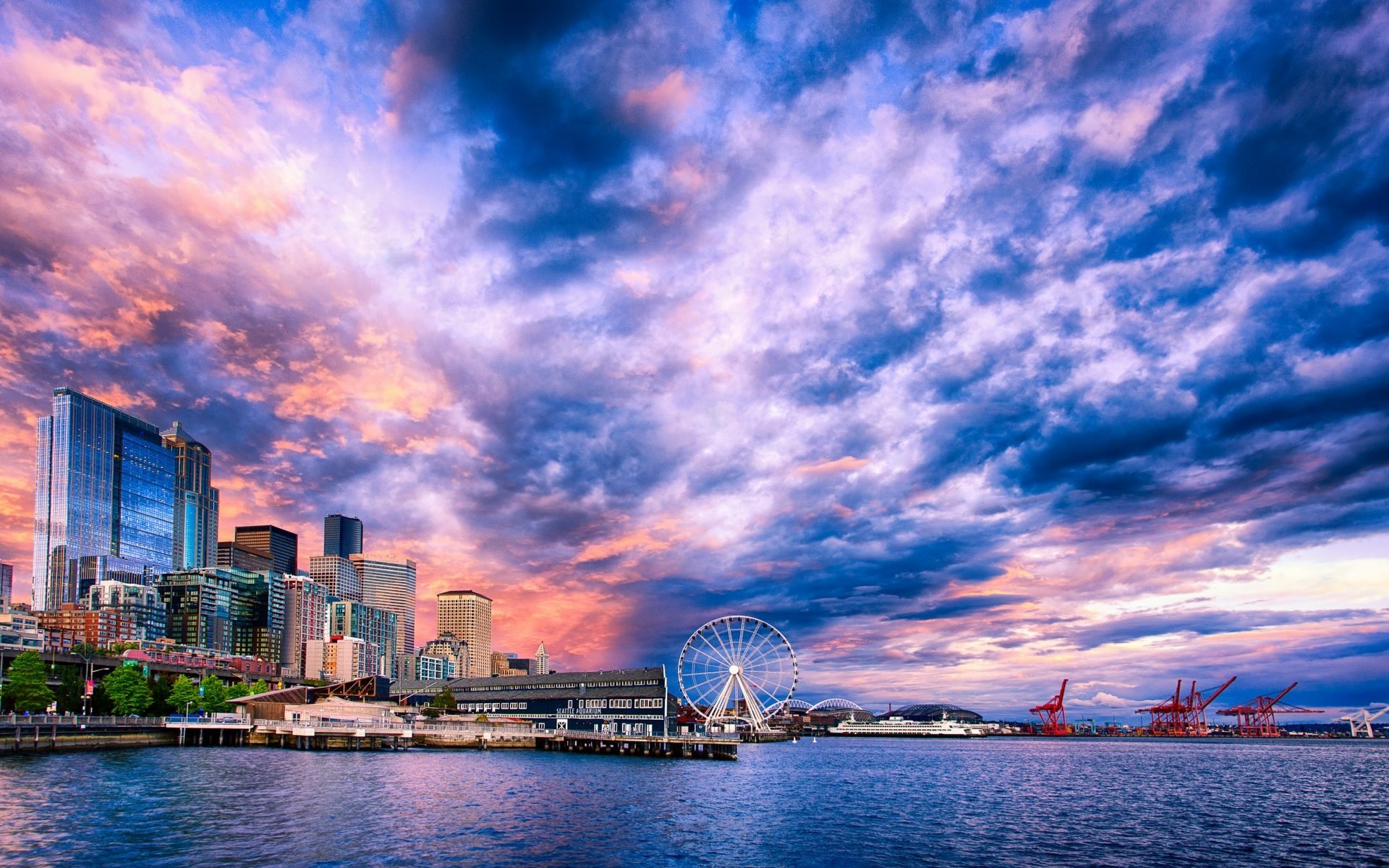 Seattle skyline at sunset with dramatic clouds over the city, featuring the waterfront, Ferris wheel, and cranes along the harbor in this HD desktop wallpaper.