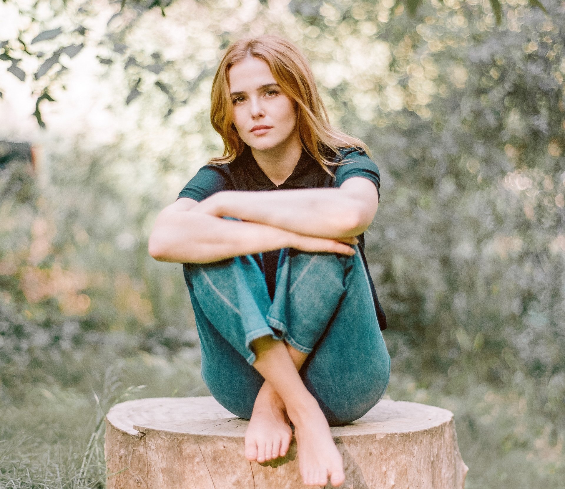 HD wallpaper of American actress Zoey Deutch with blonde hair, sitting outdoors against a soft bokeh background.