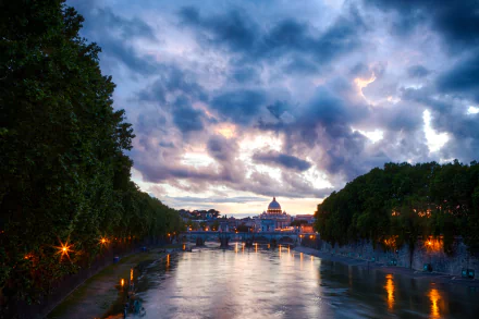 Evening clouds over the Tiber River in Rome, Italy, with city lights reflecting on the water and historic architecture visible under a dramatic sky.