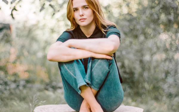 HD wallpaper of American actress Zoey Deutch with blonde hair, sitting outdoors against a soft bokeh background.