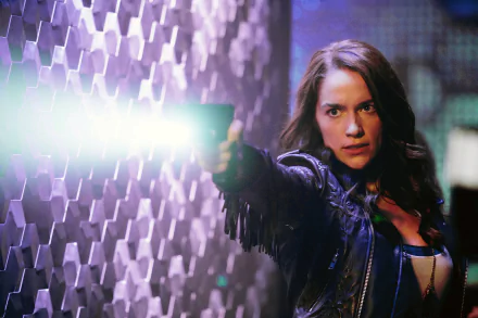 Wynonna Earp 2K desktop wallpaper: intense woman in a leather jacket aims a glowing gun against a purple-lit hexagonal wall background.
