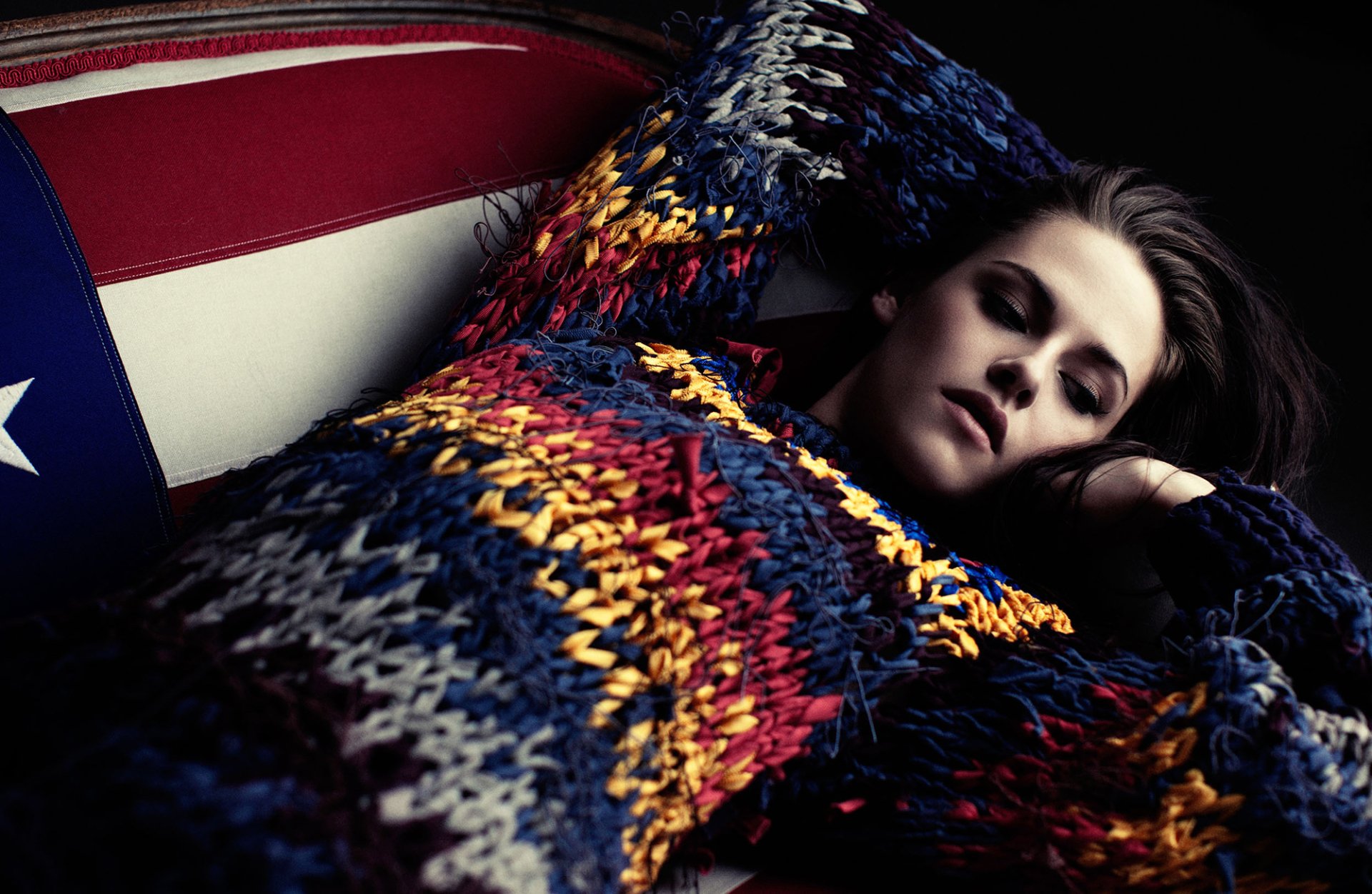 Brunette actress Kristen Stewart lounges in a colorful knitted sweater against a backdrop featuring an American flag, captured in a striking HD wallpaper style.