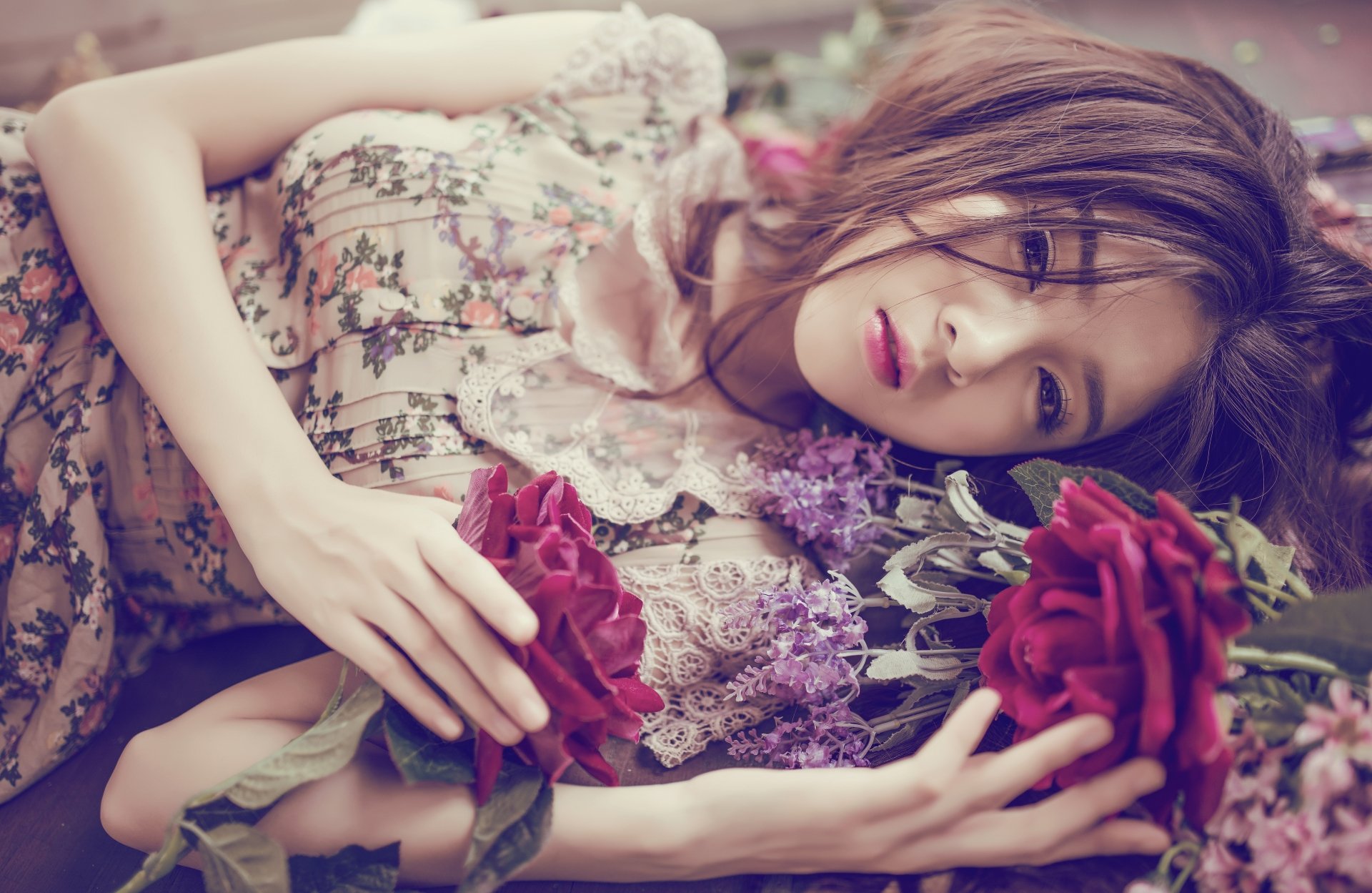 An Asian woman with brown eyes and brunette hair lies down in a flower-adorned dress, holding a red rose. This HD desktop wallpaper captures her serene and captivating beauty surrounded by flowers.