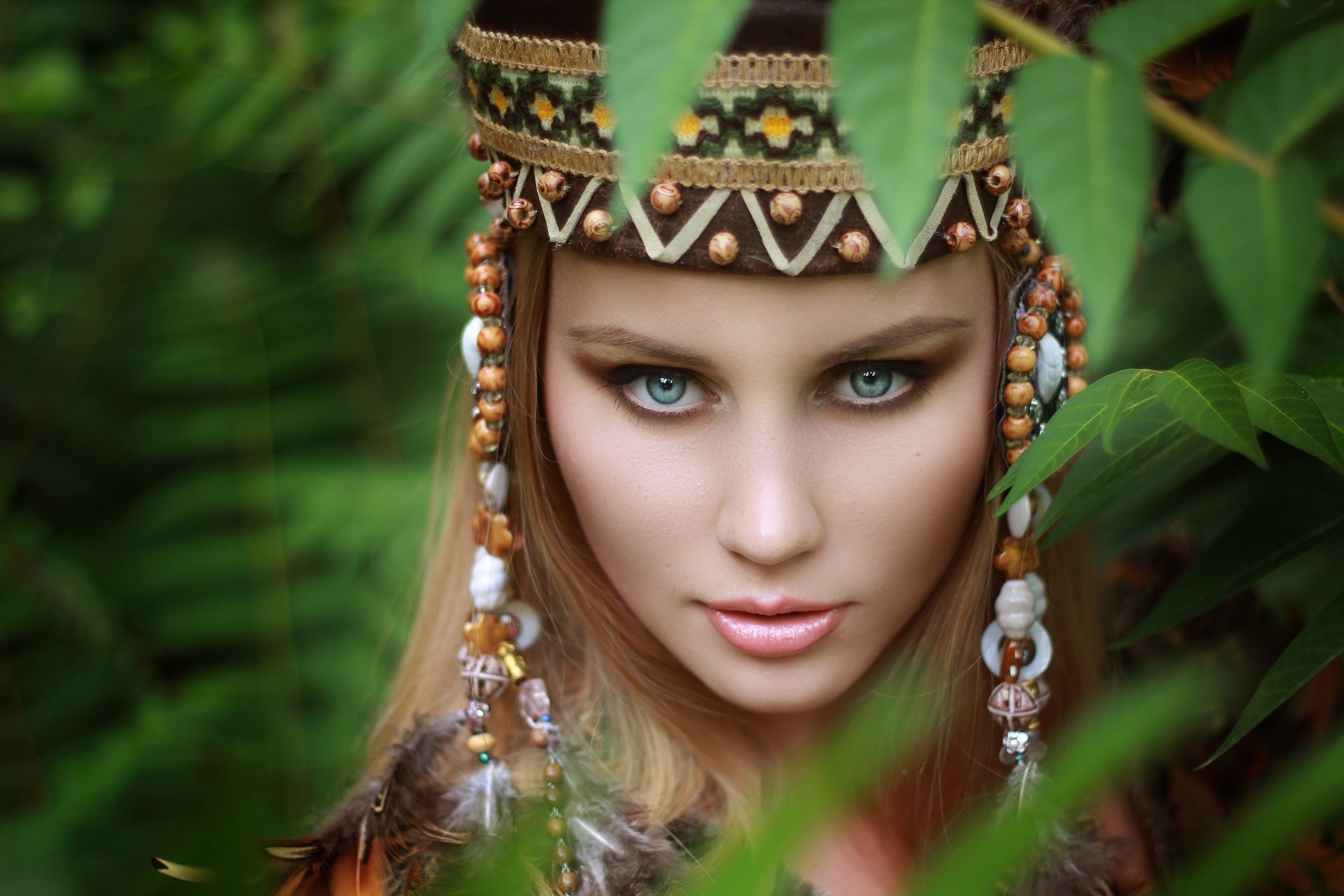 HD desktop wallpaper featuring a beautiful woman with striking blue eyes, adorned in intricate tribal jewelry, framed by lush green foliage.