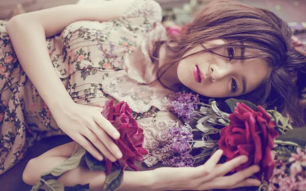 An Asian woman with brown eyes and brunette hair lies down in a flower-adorned dress, holding a red rose. This HD desktop wallpaper captures her serene and captivating beauty surrounded by flowers.