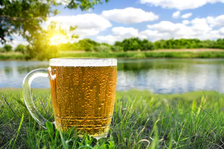 A glass of cold beer with condensation sits on grass by a lake under a bright sky, captured in stunning 4K Ultra HD for a vibrant PC desktop wallpaper.