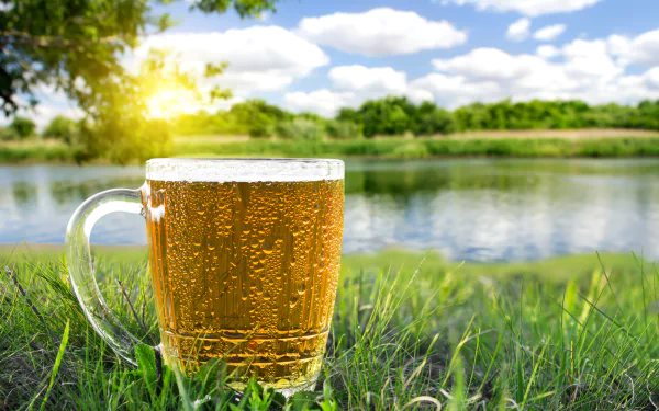 A glass of cold beer with condensation sits on grass by a lake under a bright sky, captured in stunning 4K Ultra HD for a vibrant PC desktop wallpaper.
