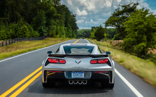 Rear view of a silver Chevrolet Corvette (C7) driving on a two-lane road surrounded by trees under a partly cloudy sky, captured as an HD PC desktop wallpaper.