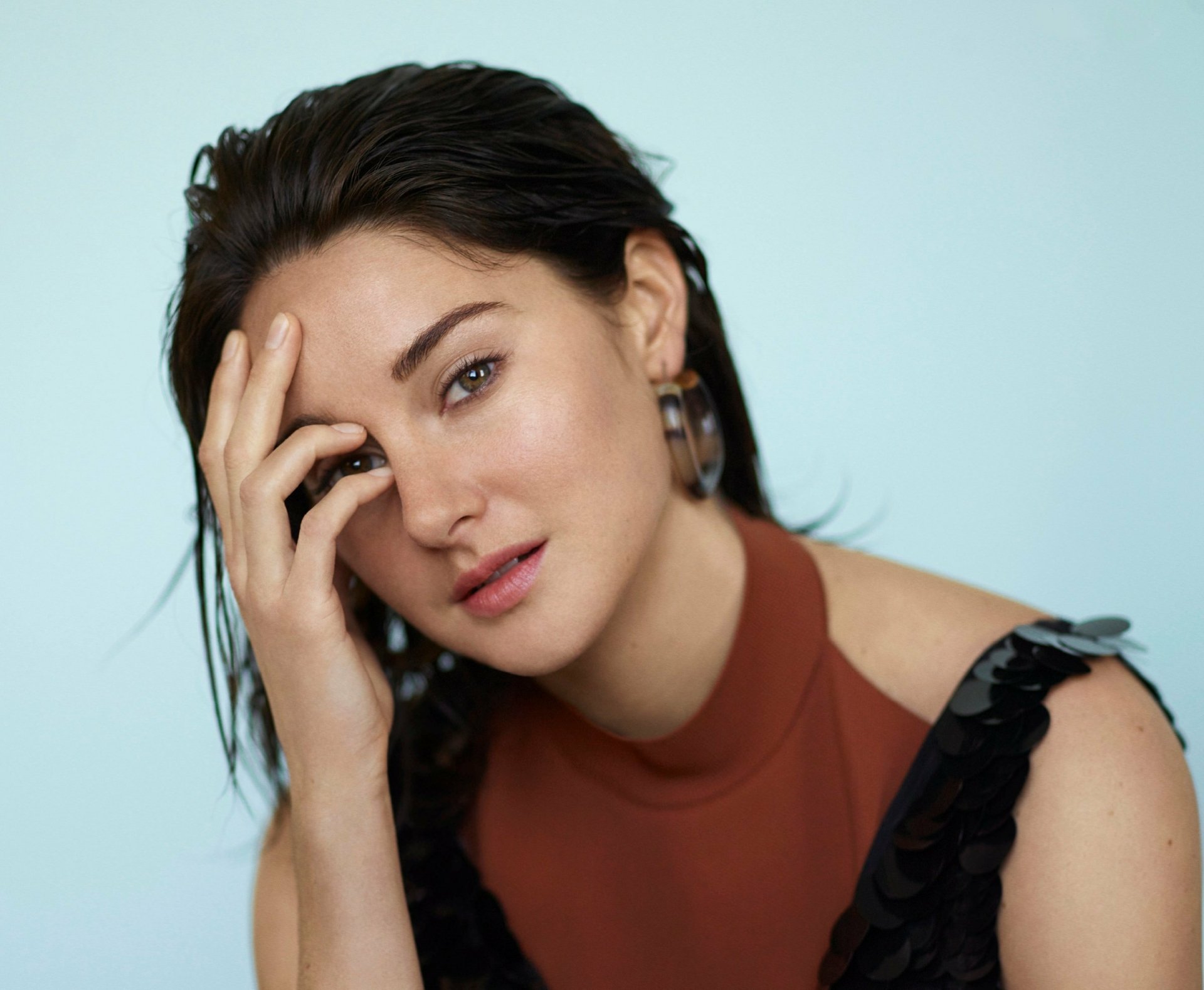 A close-up of American actress Shailene Woodley, showcasing her hazel eyes and brunette hair, set against a soft blue background, exuding natural beauty and elegance.