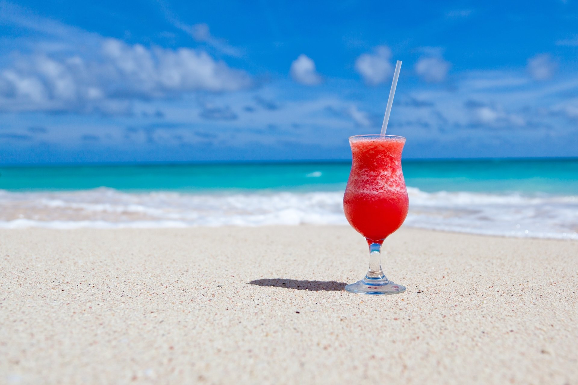A vibrant red cocktail in a glass rests on white sand at a sunny beach with blue ocean waves and sky, captured in 4K Ultra HD for a desktop wallpaper background.