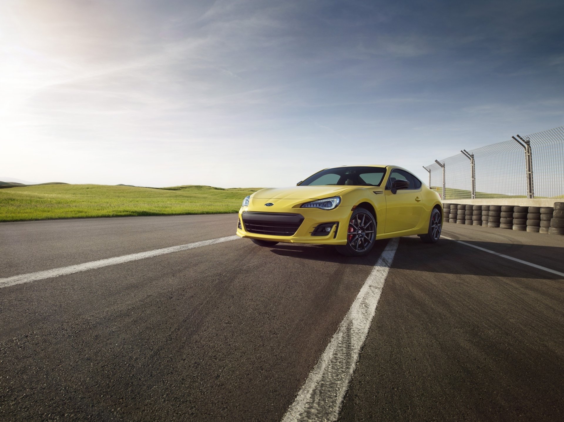 A vibrant yellow Subaru BRZ parked on a racetrack, showcasing its sleek design against a backdrop of green fields and a clear sky. A striking HD wallpaper for car enthusiasts.