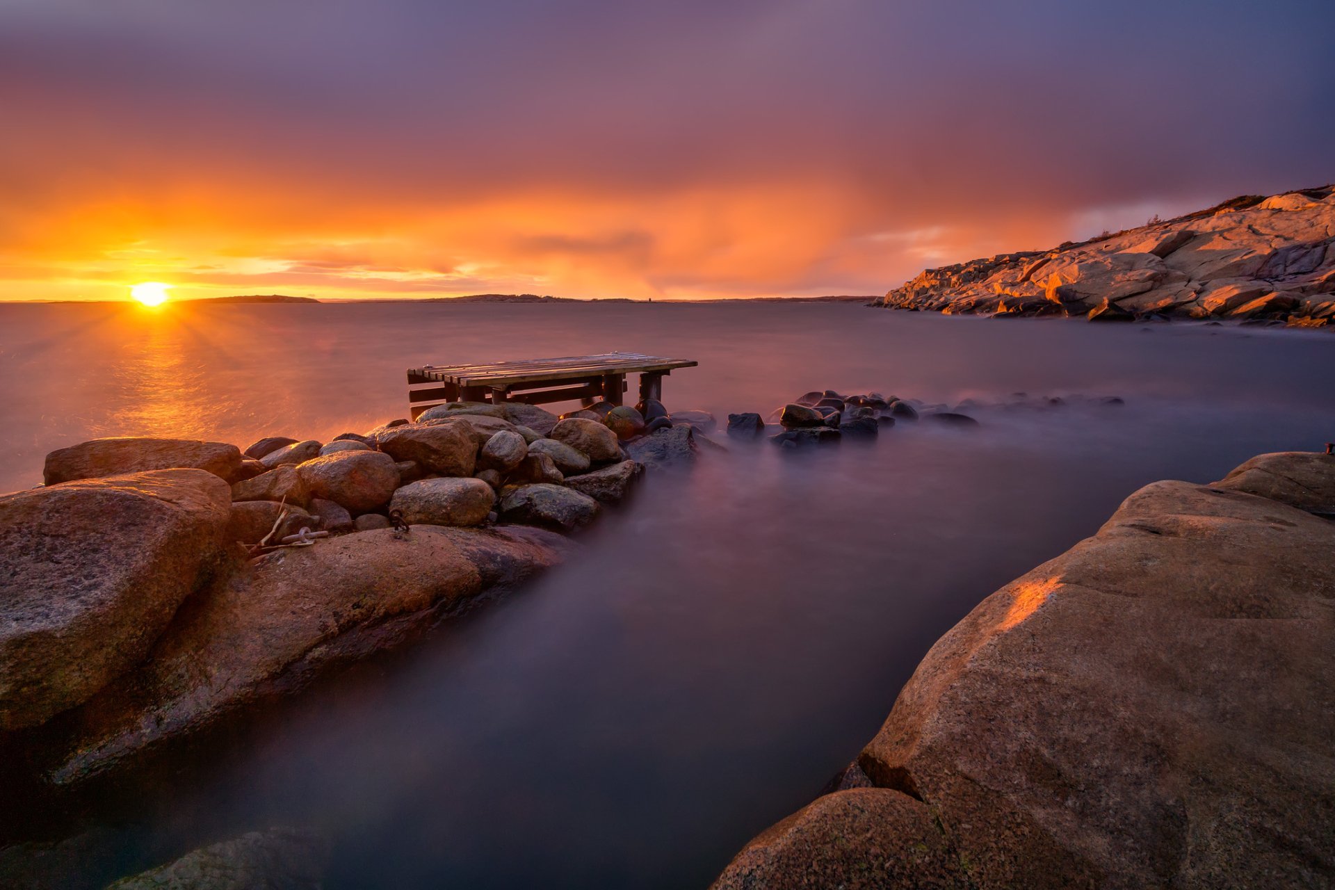 A serene sunrise over the ocean, with sunbeams illuminating the horizon. A wooden dock rests among smooth rocks, capturing the essence of nature's beauty in HD resolution.