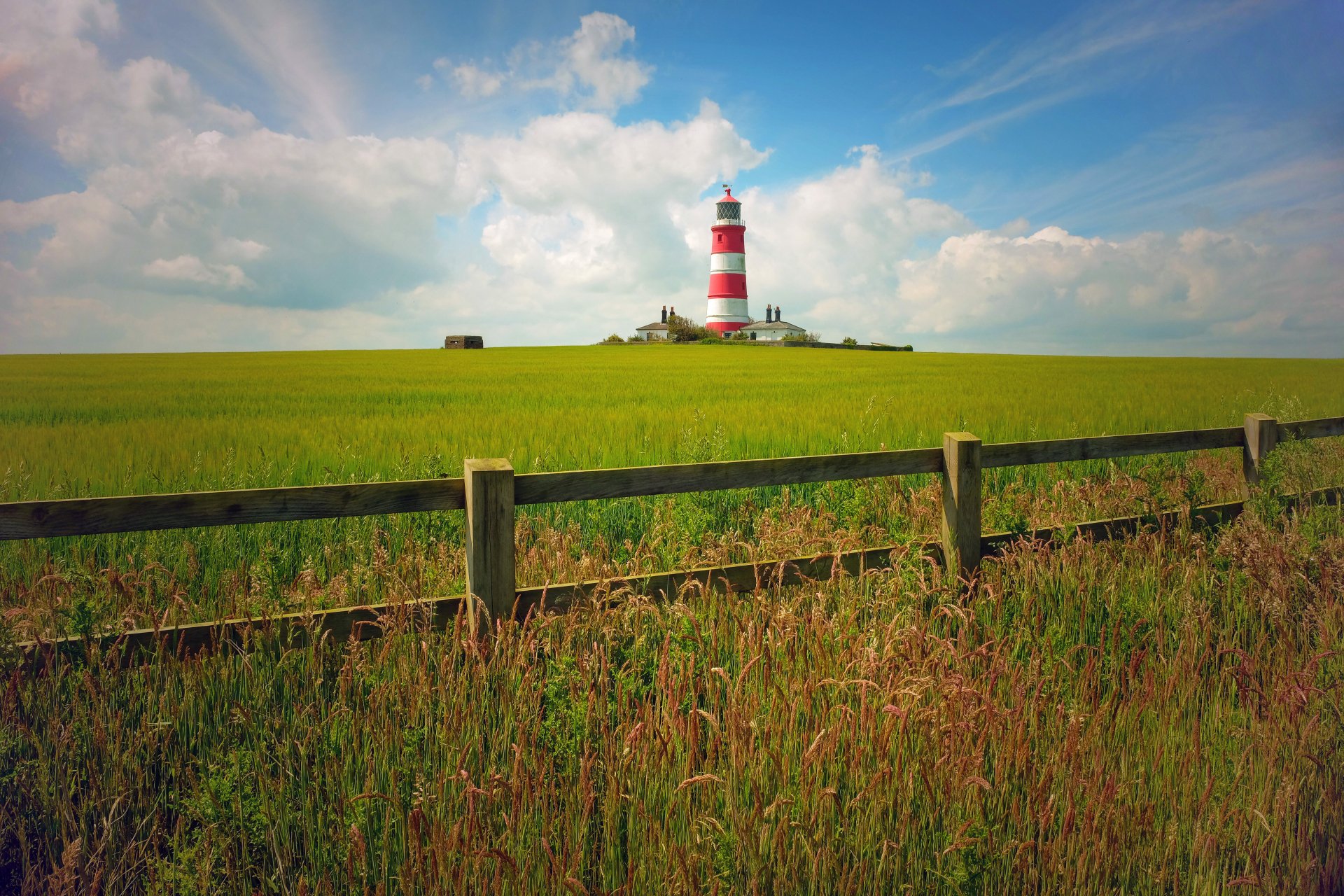 5K Ultra HD PC desktop wallpaper: red-and-white striped man-made lighthouse on a grassy hill behind a wooden fence under a blue sky with scattered clouds.