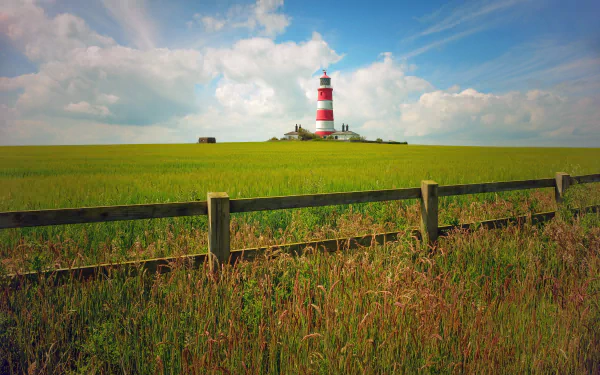 5K Ultra HD PC desktop wallpaper: red-and-white striped man-made lighthouse on a grassy hill behind a wooden fence under a blue sky with scattered clouds.