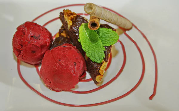 A vibrant dessert plate featuring red ice cream scoops, a biscuit topped with mint leaves and a rolled wafer, artfully presented on a white plate with a spiral sauce design in 4K Ultra HD.