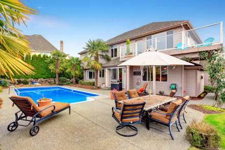 A vibrant backyard features a pool, surrounded by a stylish patio and comfortable chairs, set against a charming house, perfect for a relaxing outdoor escape.