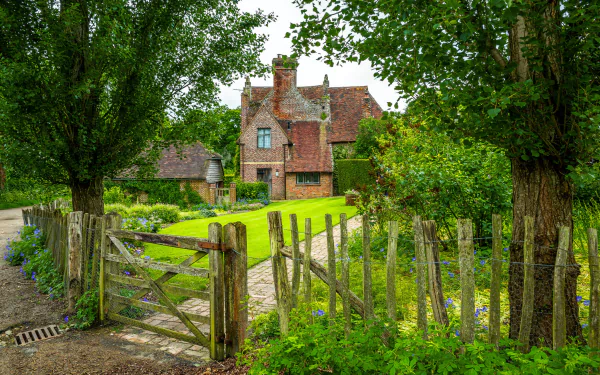 A charming English house with a brick exterior, framed by trees and a rustic wooden fence, set in a lush garden, creating a picturesque scene for an HD desktop wallpaper.