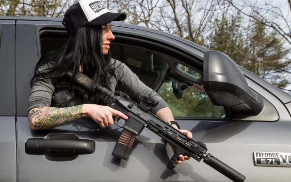 A brunette woman with tattoos holds an assault rifle while leaning out of a vehicle, wearing a cap, embodying a strong Girls & Guns aesthetic in this captivating 4K Ultra HD wallpaper.