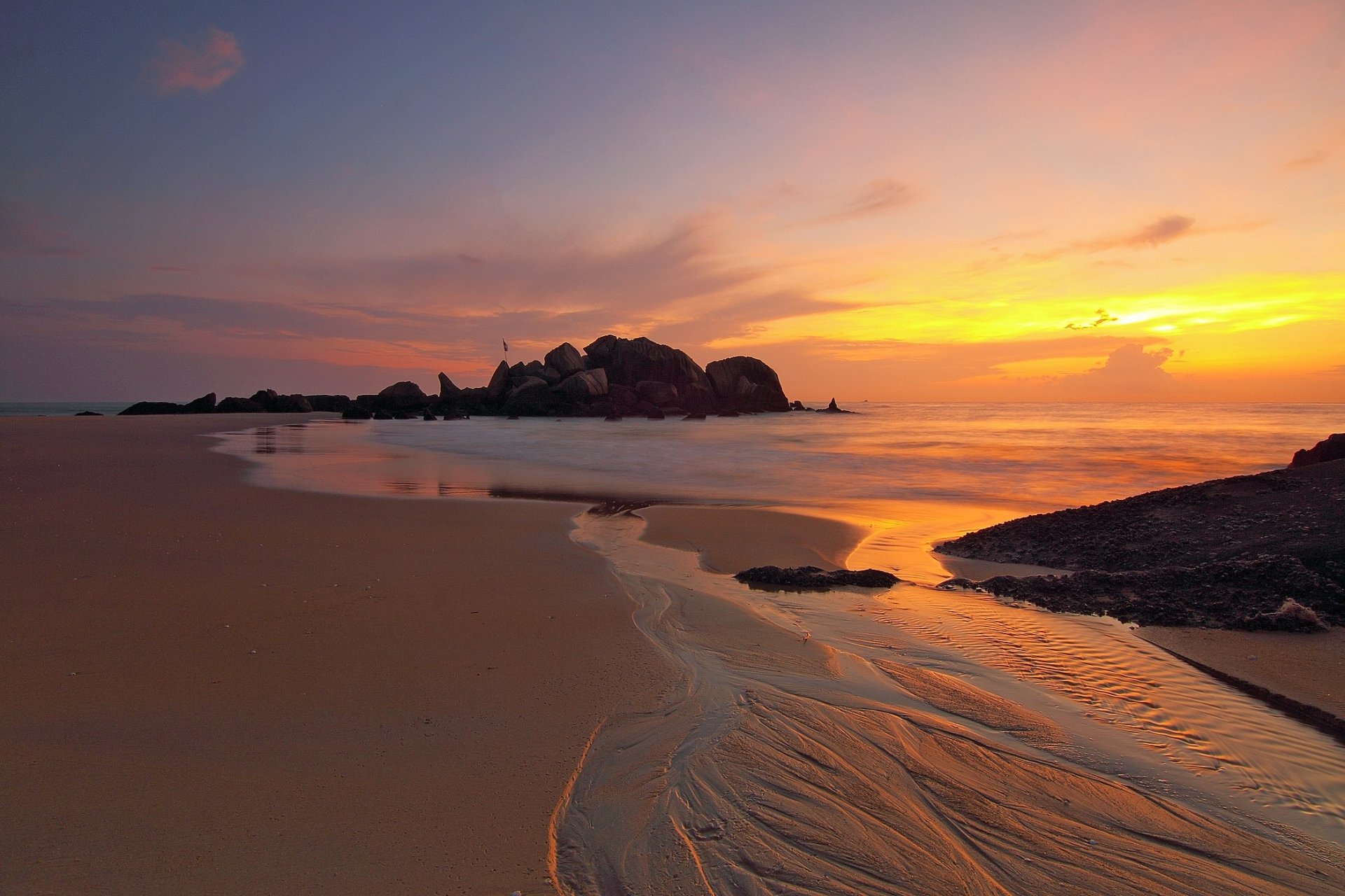 A serene seascape at sunrise, featuring an orange glow over the ocean, gently lapping waves on the sand, and rocky formations on the horizon. An inspiring natural backdrop.